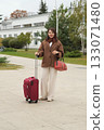 A woman in a brown wool coat stands outdoors with a suitcase, ready for travel. The frame captures movement, seasonal fashion, and calm anticipation before the journey. 133071480