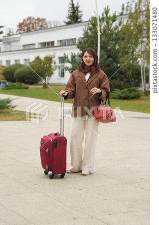 A woman in a brown wool coat stands outdoors with a suitcase, ready for travel. The frame captures movement, seasonal fashion, and calm anticipation before the journey. A woman in a brown wool coat stands outdoors with a suitcase, ready for travel. The frame captures movement, seasonal fashion, and calm anticipation before the journey. 133071480
