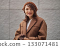 A woman in a brown coat and knitted hat smiles softly outdoors, standing against a textured wall. The image conveys autumn warmth, calm confidence, and natural elegance. 133071481