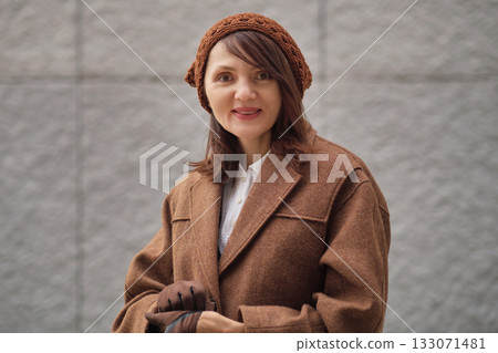 A woman in a brown coat and knitted hat smiles softly outdoors, standing against a textured wall. The image conveys autumn warmth, calm confidence, and natural elegance. A woman in a brown coat and knitted hat smiles softly outdoors, standing against a textured wall. The image conveys autumn warmth, calm confidence, and natural elegance. 133071481