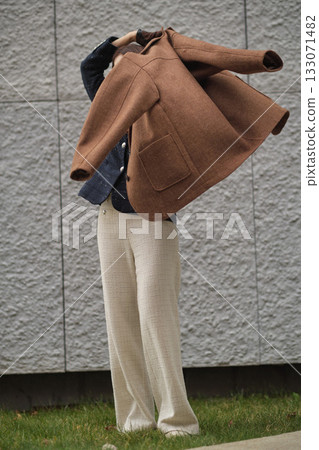 A woman lifts a brown wool coat to put it on, captured in motion against a modern gray wall. The frame reflects autumn warmth, everyday elegance, and timeless fashion in movement. A woman lifts a brown wool coat to put it on, captured in motion against a modern gray wall. The frame reflects autumn warmth, everyday elegance, and timeless fashion in movement. 133071482