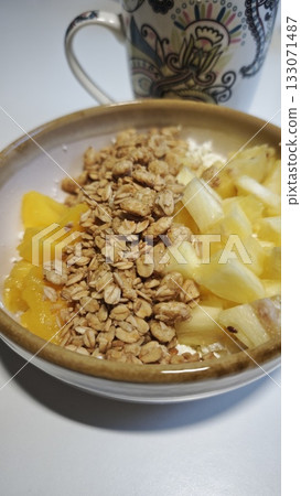 Bowl of granola with pineapple and peach pieces served with a patterned cup on a white table. The image represents healthy eating, freshness and balanced morning nutrition Bowl of granola with pineapple and peach pieces served with a patterned cup on a white table. The image represents healthy eating, freshness and balanced morning nutrition 133071487