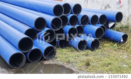 Stack of large blue plastic pipes lying outdoors on wet grass near a wall. The image represents construction, industry and infrastructure development Stack of large blue plastic pipes lying outdoors on wet grass near a wall. The image represents construction, industry and infrastructure development 133071492