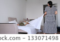 A woman laughs mid motion as she flips a crisp sheet across the bed while her son relaxes, photographed with clean composition. Daily habits shape care, hygiene, and trust in modern family life. 133071498
