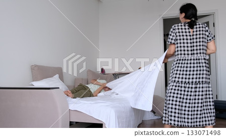 A woman laughs mid motion as she flips a crisp sheet across the bed while her son relaxes, photographed with clean composition. Daily habits shape care, hygiene, and trust in modern family life. A woman laughs mid motion as she flips a crisp sheet across the bed while her son relaxes, photographed with clean composition. Daily habits shape care, hygiene, and trust in modern family life. 133071498