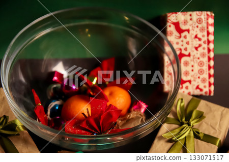 Glass bowl with Christmas ornaments, poinsettias, tangerines on dark table, wrapped gifts in kraft paper beside. Top view. Low key Glass bowl with Christmas ornaments, poinsettias, tangerines on dark table, wrapped gifts in kraft paper beside. Top view. Low key 133071517