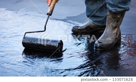 Applying a protective black coating on a flat surface with a roller tool, using work boots and blue jeans in outdoor setting, ensuring waterproofing and durability. 133071628