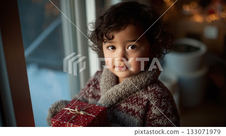 A beautiful toddler, adorned in a cozy winter sweater, holds a red gift box with a golden ribbon, creating a heartwarming festive moment by the bright window, waiting for holiday. 133071979