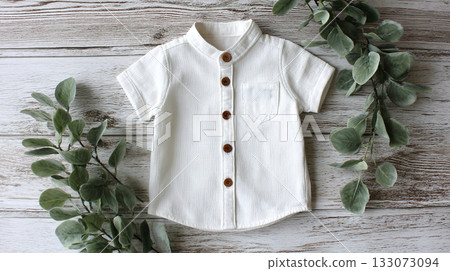 Elegant child's white shirt, short-sleeved, adorned with wood buttons, on rustic wood backdrop, complemented by green foliage. Pure, minimalist style, stylish. 133073094
