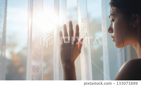 A woman's profile gazing at the light through the curtains. The soft backlighting conveys a sense of tranquility and sadness. A woman's profile gazing at the light through the curtains. The soft backlighting conveys a sense of tranquility and sadness. 133073899