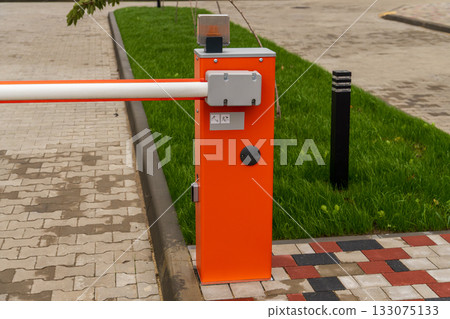 A bright orange parking barrier stands next to a sidewalk, controlling access to a clean and tidy parking area with green grass nearby 133075133