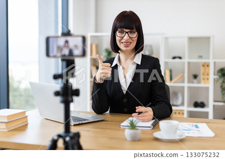 Caucasian woman wearing business attire blogging about professional topics at table with accessories. 133075232