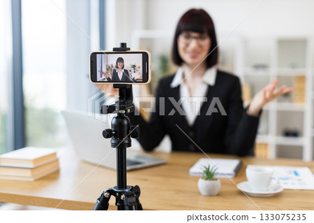 Caucasian woman in early 30s sitting at desk recording video blog about business-related topics. 133075235