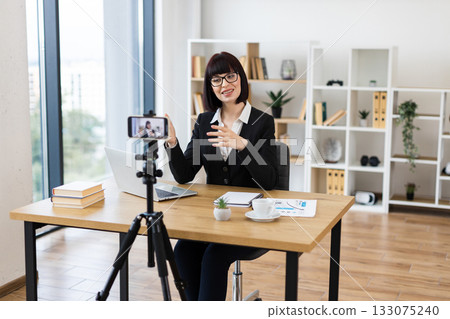 Caucasian woman sitting at desk recording video blog about business in modern office setting. Image captures professional blogging, digital marketing, technology use for strategies and ideas. Caucasian woman sitting at desk recording video blog about business in modern office setting. Image captures professional blogging, digital marketing, technology use for strategies and ideas. 133075240