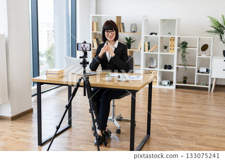 Caucasian woman sitting at desk recording video blog about business in modern office setting. Image captures professional blogging, digital marketing, technology use for strategies and ideas. Caucasian woman sitting at desk recording video blog about business in modern office setting. Image captures professional blogging, digital marketing, technology use for strategies and ideas. 133075241