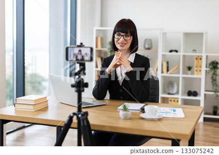 Caucasian woman wearing business attire recording video blog about professional topics using smartphone. Office setting includes desk, laptop, coffee cup, and organized shelf in background Caucasian woman wearing business attire recording video blog about professional topics using smartphone. Office setting includes desk, laptop, coffee cup, and organized shelf in background 133075242