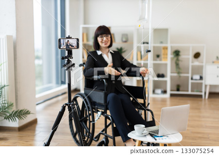 Caucasian woman in wheelchair receiving intravenous drip recording vlog about combining work with treatment using smartphone camera and laptop in modern office space 133075254