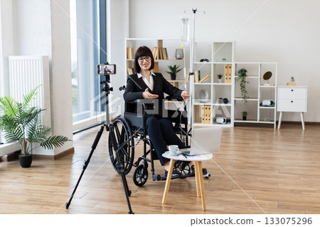 Caucasian woman in wheelchair receiving intravenous drip recording blog on camera discussing balancing work and health. Inspiring scene showcases determination, positivity, professional attire 133075296