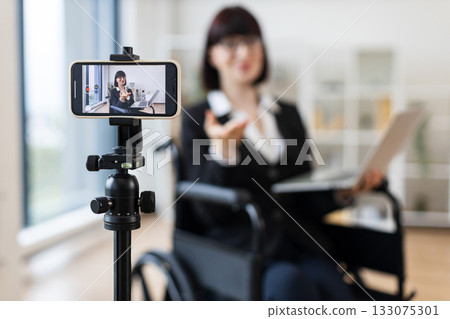 Caucasian woman in smart suit sitting in wheelchair in office during video blogging session about business practices using smartphone on tripod, promoting communication, inclusivity 133075301