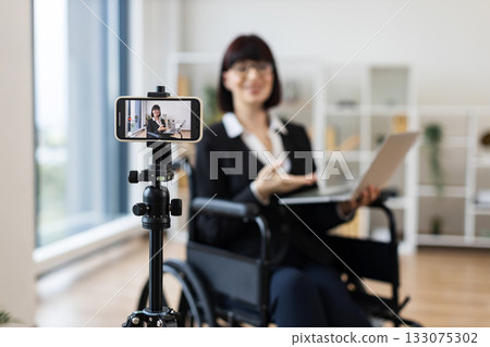 Caucasian adult woman sitting in wheelchair recording business presentation with smartphone and laptop in modern office. 133075302