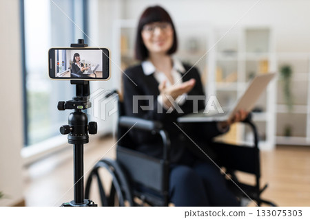 Caucasian adult woman sitting in wheelchair recording business presentation with smartphone and laptop in modern office. 133075303