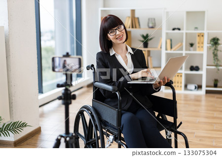 Caucasian woman in wheelchair holding laptop blogging about business using camera in office. 133075304