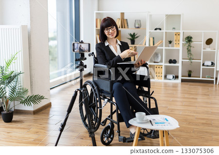 Caucasian woman in wheelchair blogs about business on camera using modern equipment. Caucasian woman in wheelchair blogs about business on camera using modern equipment. 133075306
