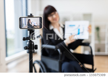 Caucasian woman wearing business attire in wheelchair records blog on smartphone tripod about business topics. Scene emphasizes professionalism, adaptability, modern business approach. Caucasian woman wearing business attire in wheelchair records blog on smartphone tripod about business topics. Scene emphasizes professionalism, adaptability, modern business approach. 133075309