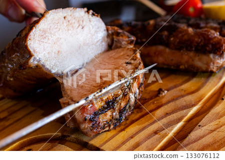 Hand cutting thick slices of grilled beef picanha off the bone on a wooden cutting board, 133076112