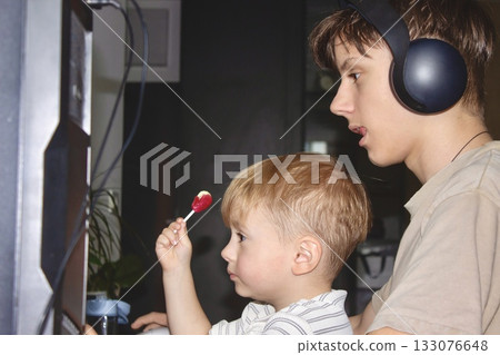 Teenager wearing headphones holding a little boy with lollipop while playing computer games. Teenager wearing headphones holding a little boy with lollipop while playing computer games. 133076648