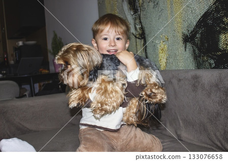 A cheerful little boy is playing and holding a small Yorkshire Terrier dog in his hands. The child looks joyful and affectionate, expressing love for pets and the bond between kids and animals. 133076658
