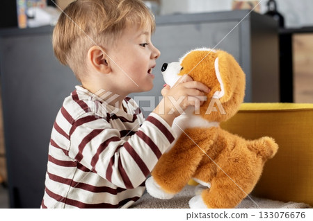 A cheerful little boy plays with a plush toy dog at home. The child is smiling and engaged in imaginative play, expressing emotions and curiosity. Cozy indoor atmosphere. A cheerful little boy plays with a plush toy dog at home. The child is smiling and engaged in imaginative play, expressing emotions and curiosity. Cozy indoor atmosphere. 133076676