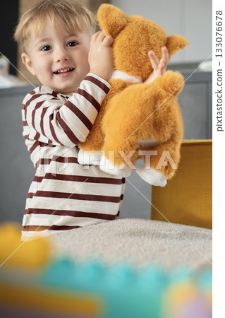 A cheerful little boy plays with a plush toy dog at home. The child is smiling and engaged in imaginative play, expressing emotions and curiosity. Cozy indoor atmosphere.  133076678