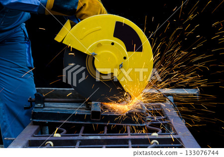 Worker in blue coveralls using yellow chop saw to cut metal material with orange sparks from cutting point 133076744
