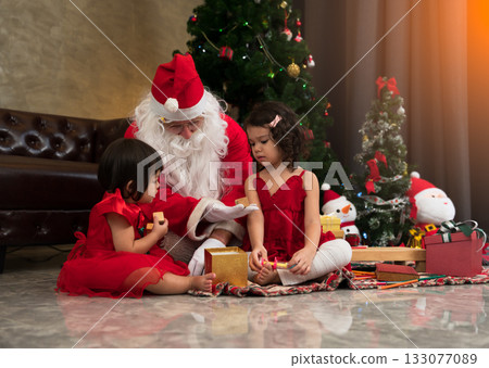 Two young girls in red dresses sitting on floor with Santa Claus beside a decorated Christmas tree while  open presents together in living room at home 133077089