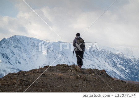 Hiker standing on rocky edge overlooking snow covered mountains in remote wilderness 133077174
