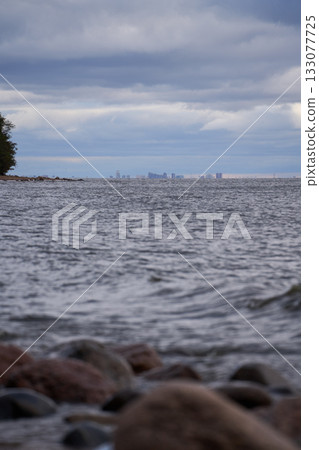 Distant city skyline over Gulf of Finland with stones and waves in foreground. 133077725