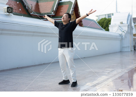 A man opens his arms wide in a quiet Bangkok temple courtyard, lifting his gaze with a feeling of calm, clarity, and emotional release. 133077859