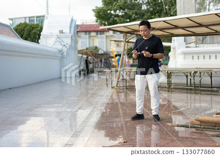 A man stands in a Bangkok temple courtyard, taking his glasses from their case as he prepares to put them on amid the calm traditional surroundings. 133077860