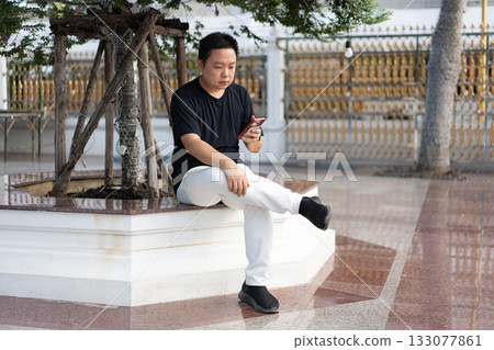A man sits in the shade beside a tree planter at a Bangkok temple, relaxed as he checks his phone in a quiet outdoor moment. 133077861