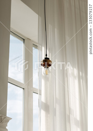 Modern hanging light fixture with soft natural light through sheer curtains and large window 133079157