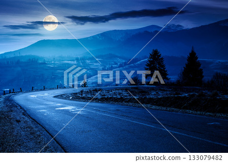country road in mountains at night. beautiful countryside landscape in late autumn. fir trees on the roadside in full moon light. scenic view of blue sky with clouds above the distant ridge in snow country road in mountains at night. beautiful countryside landscape in late autumn. fir trees on the roadside in full moon light. scenic view of blue sky with clouds above the distant ridge in snow 133079482