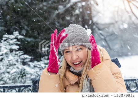 Portrait young adult smiling woman wear long quilted coat white hat enjoy play snowball snowy day outdoors. Snowflakes fall she moves playfully in front of stairs decorated with glowing winter lights 133079932