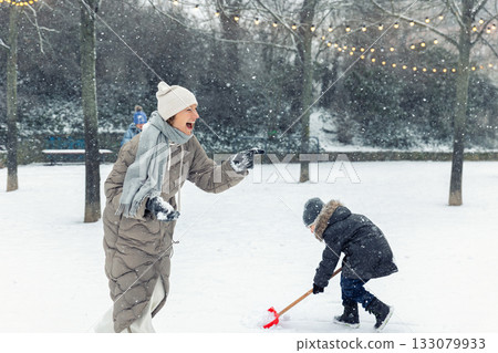Woman in a long winter coat and hat plays joyfully in the snow, laughing while engaging with a child shoveling snow nearby. Falling snow and festive lights create a cheerful winter family scen 133079933