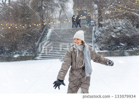 Portrait young adult smiling woman wear long quilted coat white hat enjoy play snowball snowy day outdoors. Snowflakes fall she moves playfully in front of stairs decorated with glowing winter lights Portrait young adult smiling woman wear long quilted coat white hat enjoy play snowball snowy day outdoors. Snowflakes fall she moves playfully in front of stairs decorated with glowing winter lights 133079934