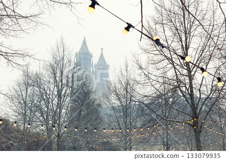 Snow falls over Magdeburg Cathedral, partly hidden behind bare winter trees and glowing garlands of yellow lights. The soft fog and quiet ambiance create a dreamy festive city atmosphere 133079935