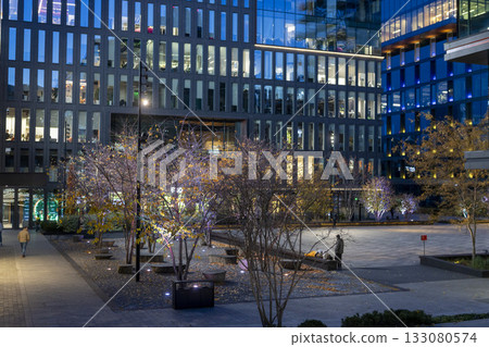 An evening view of a business center with modern glass and steel architecture and a square with a flowerbed next to it. 133080574