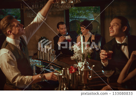 Retro-dressed people chatting at the bar and enjoying evening cocktails Retro-dressed people chatting at the bar and enjoying evening cocktails 133080833