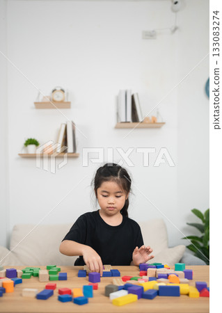 Focused girl building with colorful blocks on wooden table in modern living room, engaging in creative play, enhancing motor skills and cognitive development Focused girl building with colorful blocks on wooden table in modern living room, engaging in creative play, enhancing motor skills and cognitive development 133083274