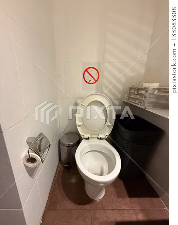 Toilet with white tiled walls, trash bin, and paper holder. Simple hygiene concept, minimal design, and clean interior detail. Toilet with white tiled walls, trash bin, and paper holder. Simple hygiene concept, minimal design, and clean interior detail. 133083308